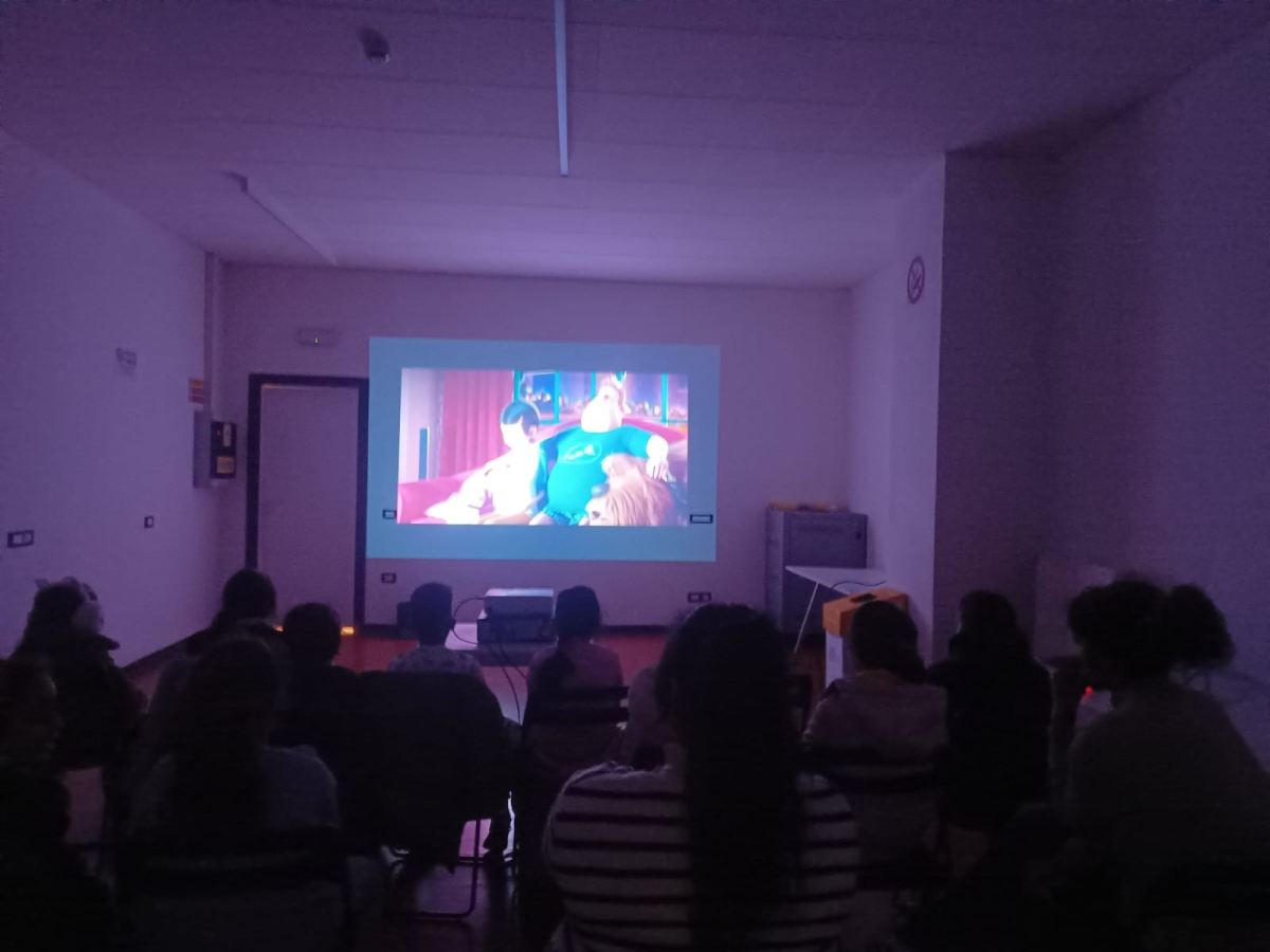 Pubblico seduto in una sala buia che guarda la proiezione di un film o video su grande schermo. 