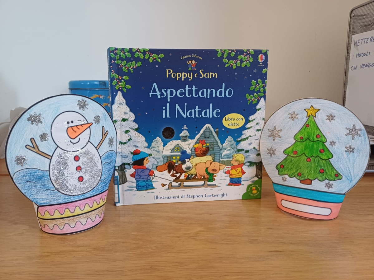 Libro per bambini &quot;Poppy e Sam Aspettando il Natale&quot; esposto centralmente tra due decorazioni artigianali a forma di palla di neve natalizia. 