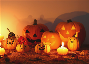 Un gruppo di zucche di Halloween intagliate e illuminate, con candele accese e foglie secche in primo piano, su uno sfondo caldo arancione. 