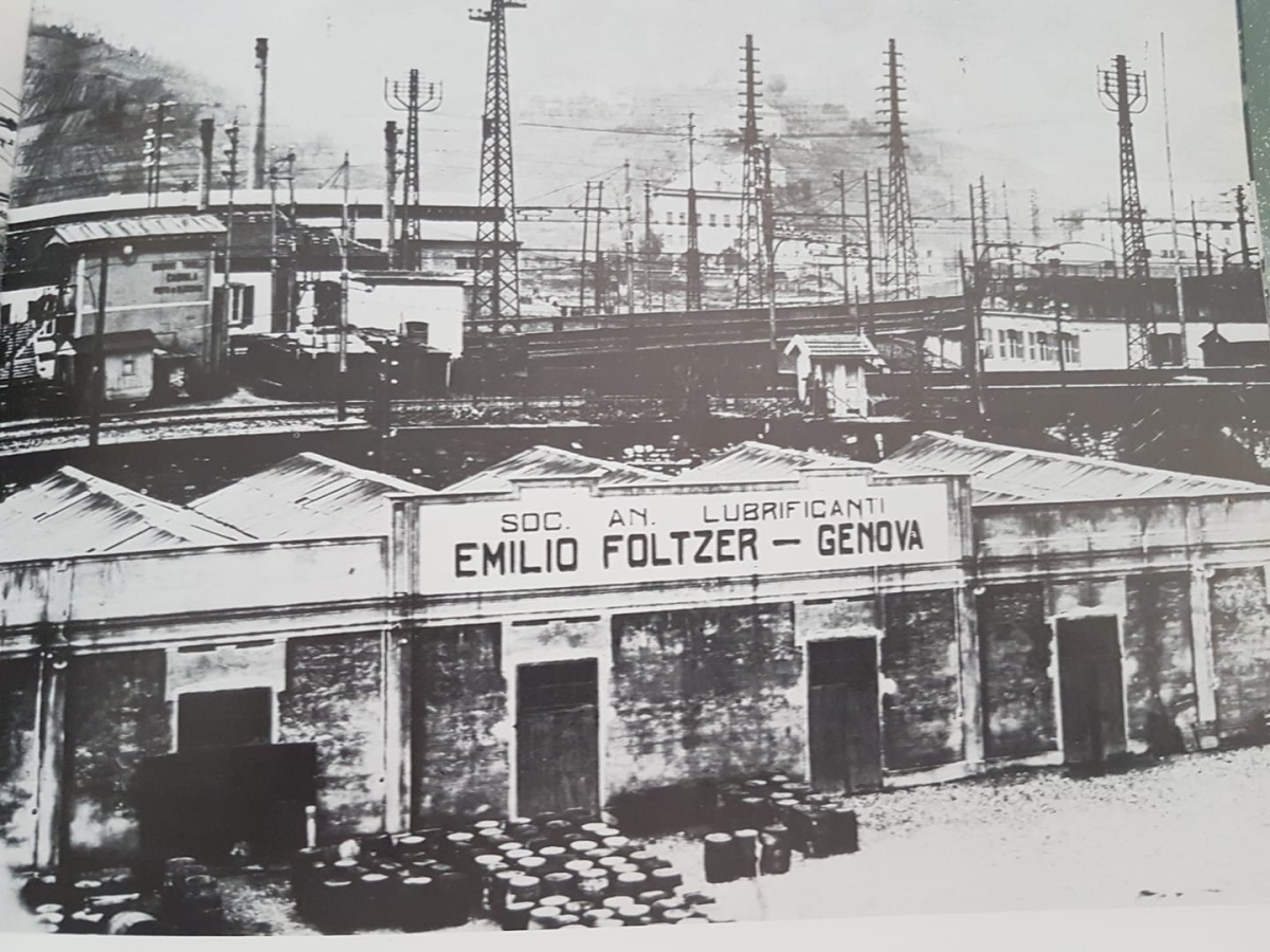 Una vecchia fotografia in bianco e nero di un magazzino industriale con l'insegna "Soc. An. Lubrificanti Emilio Foltzer – Genova". 