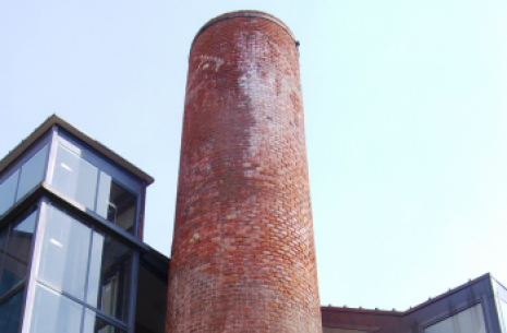 Ciminiera in mattoni rossi si erge accanto a edifici moderni con ampie vetrate.