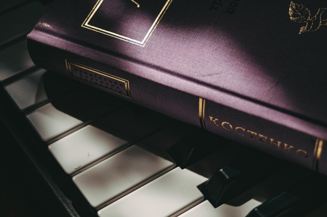 Un primo piano un libro con la copertina viola scuro e scritte dorate, poggiato sulle tastiere bianche e nere di un pianoforte.