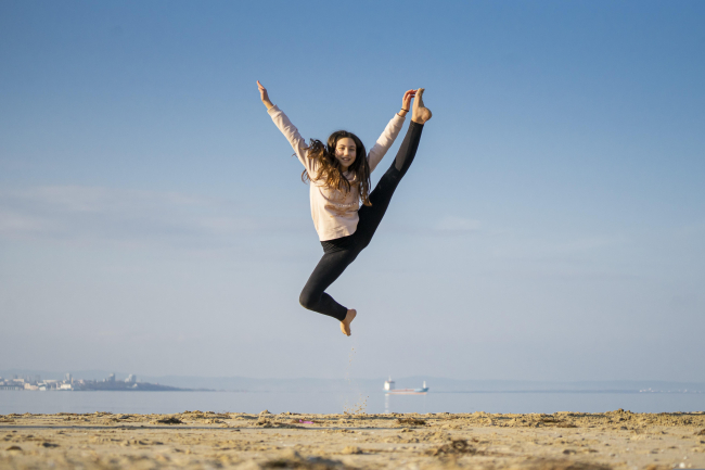Una ragazza sorridente cattura un salto acrobatico in aria, con una gamba tesa verso l'alto e le braccia aperte, su una spiaggia sabbiosa con il mare, una nave e un cielo azzurro sullo sfondo.